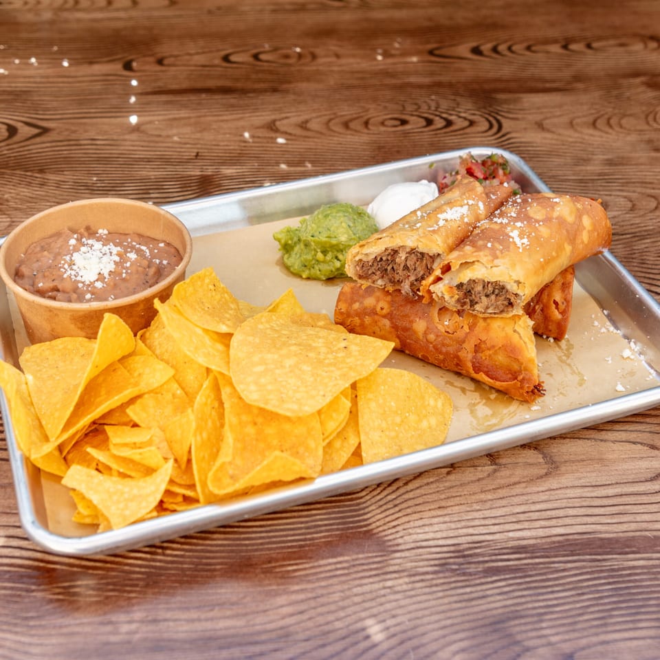 Crispy taquitos, one cut open to show meat filling, with tortilla chips, refried beans, guacamole, and sour cream.