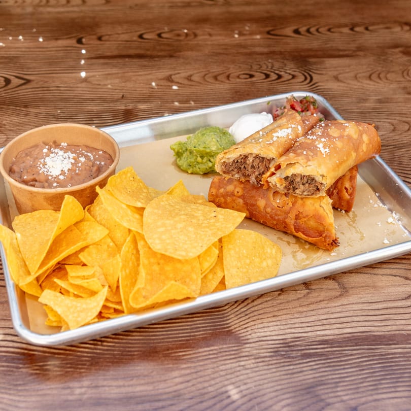 Crispy taquitos, one cut open to show meat filling, with tortilla chips, refried beans, guacamole, and sour cream.