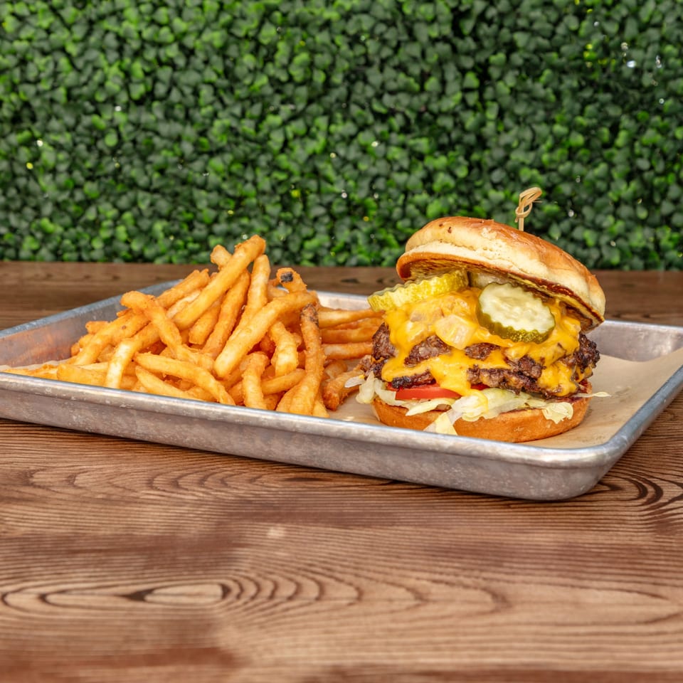 Wagyu Smashburger with melted cheese, pickles, and fries on a metal tray against a green wall.