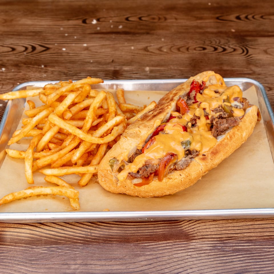 A Wagyu Smashburgers & Tacos cheesesteak with fries on a metal tray with a wooden background.