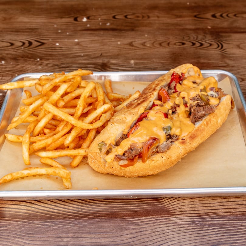 A Wagyu Smashburgers & Tacos cheesesteak with fries on a metal tray with a wooden background.