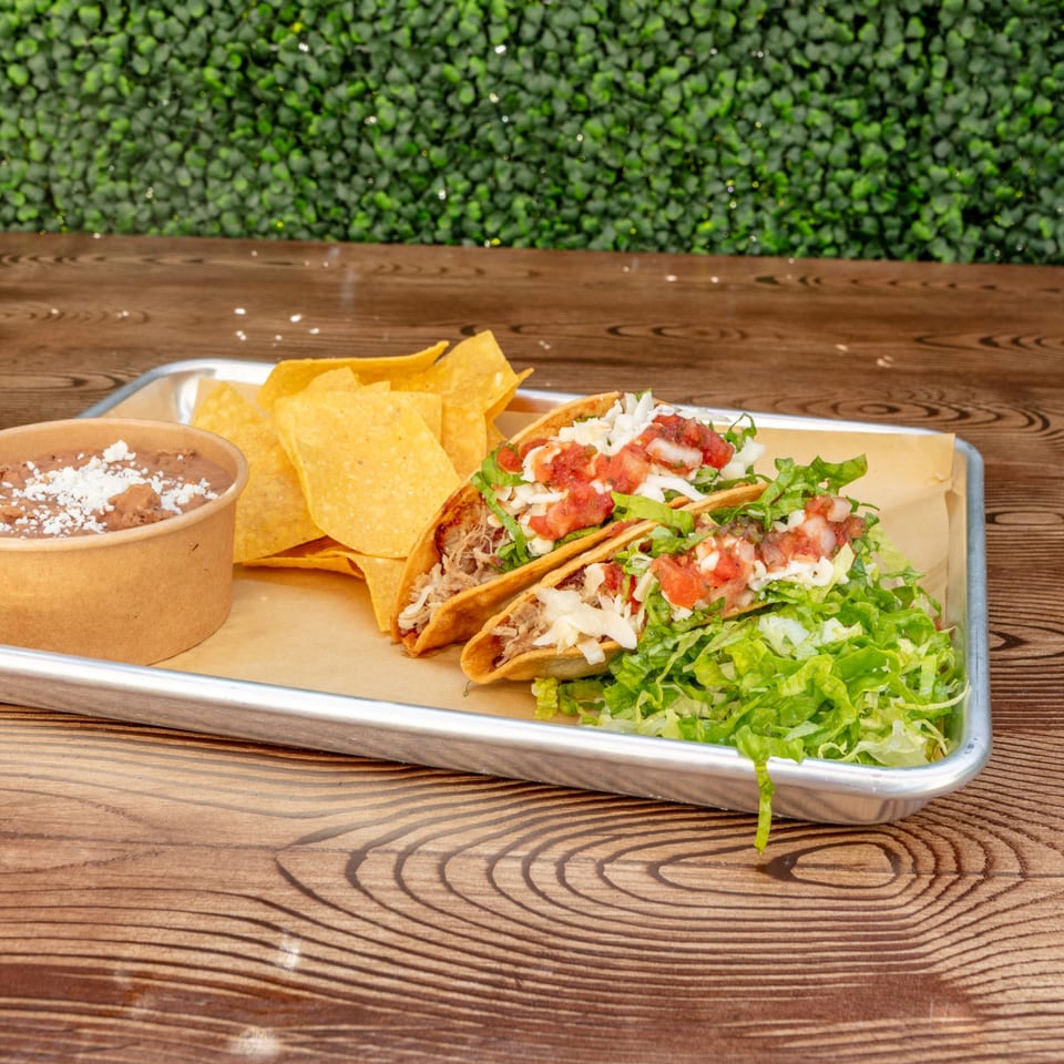 Two tacos with shredded meat, lettuce, cheese, and salsa, with chips and beans on a tray.